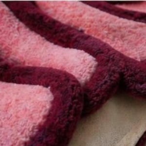 May include: Close-up of a soft, textured rug with a wavy design. The rug features alternating stripes of light pink and deep burgundy. The burgundy stripes have a raised, rounded edge, creating a three-dimensional effect. The rug's base is a light beige color.