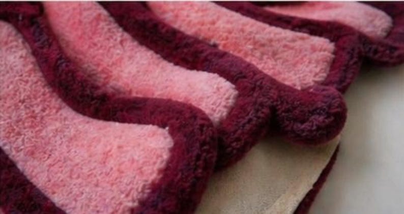 May include: Close-up of a soft, textured rug with a wavy design. The rug features alternating stripes of light pink and deep burgundy. The burgundy stripes have a raised, rounded edge, creating a three-dimensional effect. The rug's base is a light beige color.