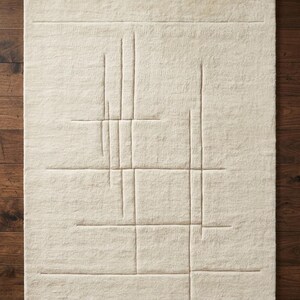 Pode incluir: Um tapete retangular creme com um design geom&eacute;trico. O tapete apresenta uma s&eacute;rie de linhas em relevo formando um padr&atilde;o semelhante a uma grade com elementos verticais e horizontais. O tapete &eacute; feito de um material macio e texturizado.