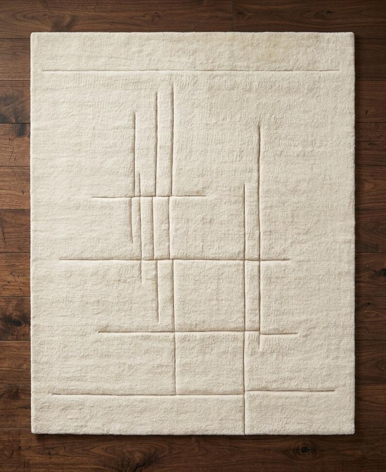 Pode incluir: Um tapete retangular creme com um design geom&eacute;trico. O tapete apresenta uma s&eacute;rie de linhas em relevo formando um padr&atilde;o semelhante a uma grade com elementos verticais e horizontais. O tapete &eacute; feito de um material macio e texturizado.