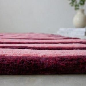 May include: A plush, striped rug in shades of pink and burgundy. The rug has a soft, textured surface with horizontal stripes. The rug is a decorative item for a living room or bedroom.