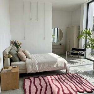 May include: A modern bedroom with a bed, pillows, and a unique rug. The rug is pink with a dark red border and an abstract design. A side table with a lamp and a chair are also visible.