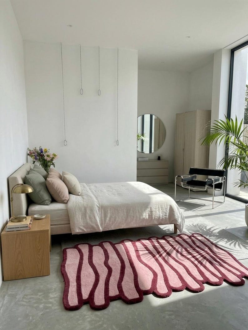 May include: A modern bedroom with a bed, pillows, and a unique rug. The rug is pink with a dark red border and an abstract design. A side table with a lamp and a chair are also visible.