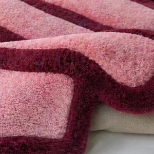 May include: Close-up of a plush, rectangular rug with a soft, textured surface. The rug features a light pink center with a deep burgundy border and stripes, creating a contrasting design. The rug is folded over, revealing a glimpse of the backing.