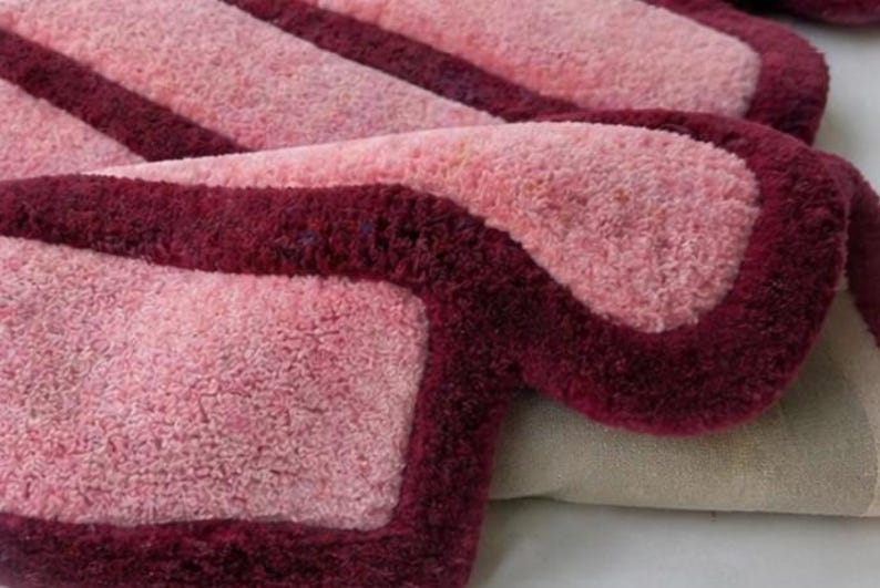 May include: Close-up of a plush, rectangular rug with a soft, textured surface. The rug features a light pink center with a deep burgundy border and stripes, creating a contrasting design. The rug is folded over, revealing a glimpse of the backing.