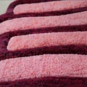 May include: Close-up of a soft, textured bath mat with a repeating pattern. The mat features alternating stripes of light pink and deep burgundy. The edges are rounded, creating a unique shape.