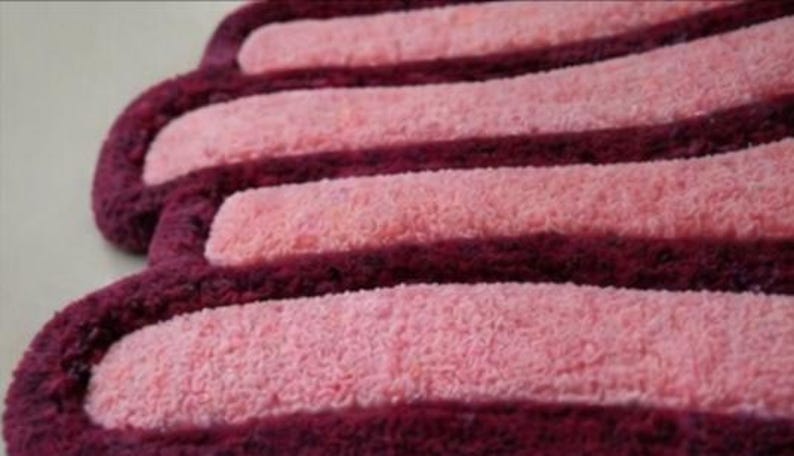 May include: Close-up of a soft, textured bath mat with a repeating pattern. The mat features alternating stripes of light pink and deep burgundy. The edges are rounded, creating a unique shape.