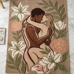 Pode incluir: Um tapete retangular tufado em cor base bronzeada com um gráfico central de duas figuras a abraçar-se e a beijar-se. O design é rodeado por lírios brancos, folhas verdes e rosas de cor pêssego suave, tudo em pelo texturizado.