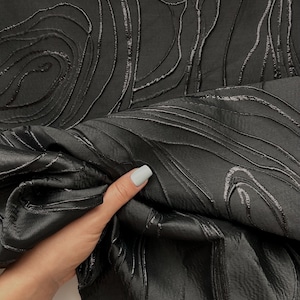 Può includere: Un tessuto nero caratterizzato da un motivo strutturato in rilievo con linee organiche vorticose che creano un effetto topografico. Il materiale presenta una lucentezza sottile e le linee in rilievo sono più scure della base del tessuto.