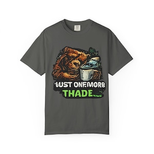 May include: Dark gray t-shirt with a graphic featuring a bear, a cup, and the text "Just One More Thade." The design includes brown, white, green, and blue elements. The shirt is a casual wear item.