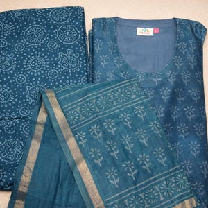 May include: A teal-colored Indian salwar kameez set. The set includes a top, bottoms, and a scarf. The top has a scoop neckline and a floral pattern. The bottoms have a circular pattern. The scarf has a floral border with gold trim.