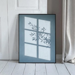 Könnte beinhalten: Ein rechteckiger Wandkunstdruck in einem dünnen schwarzen Rahmen mit einem gedämpften blauen Hintergrund. Das Bild zeigt den Schatten eines Fenstersprossengitters und die Silhouette von blättrigen Pflanzenzweigen auf der blauen Oberfläche.