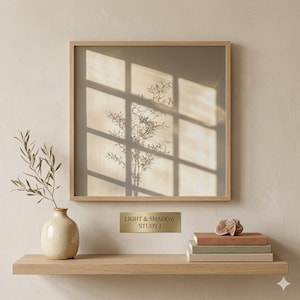 Op de afbeelding: Een ingelijst kunstwerk met een licht houten frame, met de schaduw van een plant tegen een beige achtergrond. Daaronder een plank met een vaas, boeken en een bordje met "LIGHT & SHADOW STUDY 1". De stijl is minimalistisch en natuurlijk.