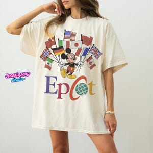 May include: Cream-colored oversized t-shirt featuring a graphic of Mickey Mouse holding flags from various countries. The word "Epcot" is printed in colorful letters below the graphic, with a globe graphic in the center. The shirt has short sleeves.