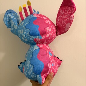 May include: A plush toy in the shape of a character with blue and pink sections. The blue sections have a white floral pattern. The pink sections have a white floral pattern. The toy has three yellow and red candle-like protrusions.