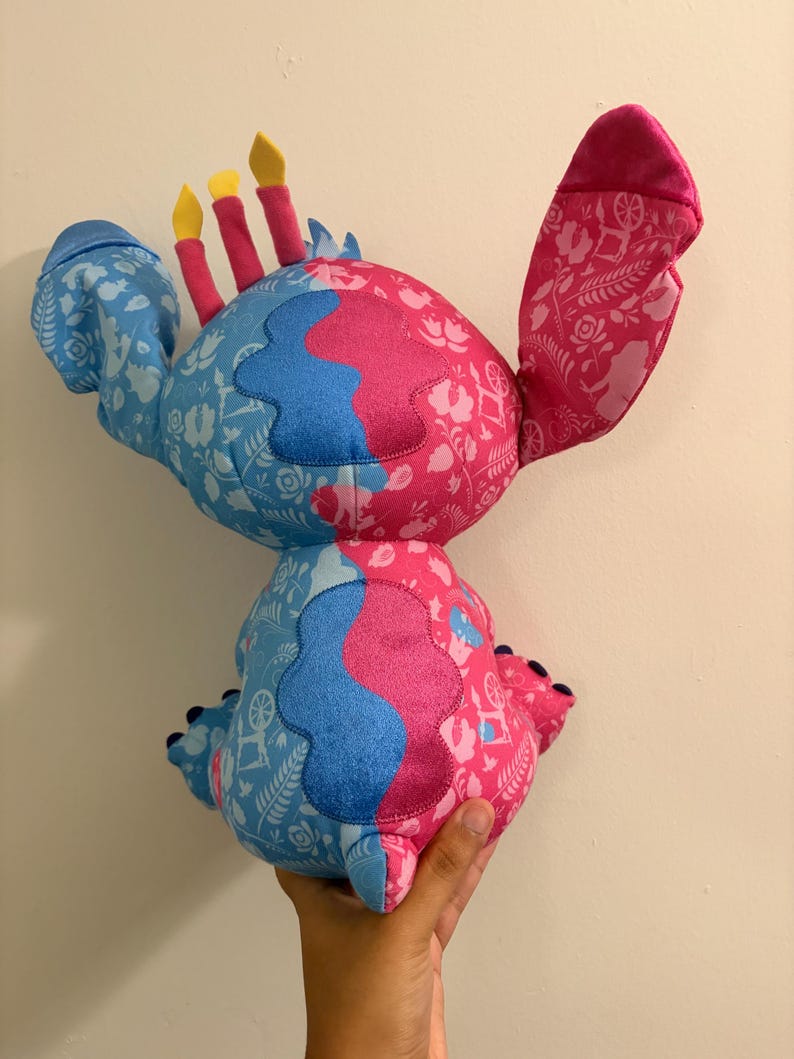 May include: A plush toy in the shape of a character with blue and pink sections. The blue sections have a white floral pattern. The pink sections have a white floral pattern. The toy has three yellow and red candle-like protrusions.