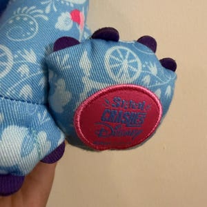 May include: Close-up of a blue plush toy foot with purple claws. The foot is decorated with a white pattern and a pink and red circular patch that reads "A Stitch Crushes Disney".