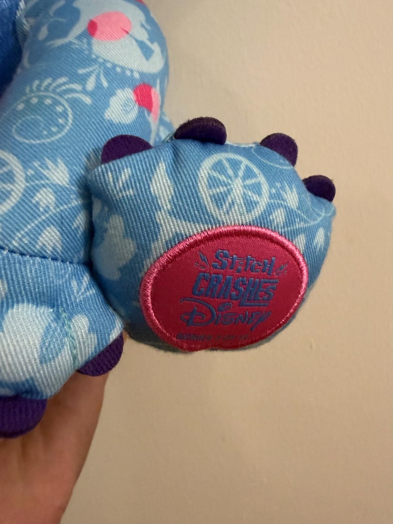 May include: Close-up of a blue plush toy foot with purple claws. The foot is decorated with a white pattern and a pink and red circular patch that reads "A Stitch Crushes Disney".