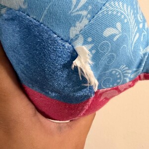 May include: Close-up of a plush toy with a tear. The toy features a combination of blue and pink fabrics. The blue fabric has a floral pattern, while the pink fabric is solid. The tear reveals the white stuffing.