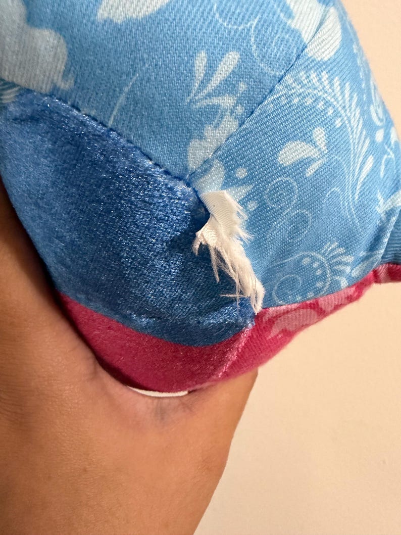 May include: Close-up of a plush toy with a tear. The toy features a combination of blue and pink fabrics. The blue fabric has a floral pattern, while the pink fabric is solid. The tear reveals the white stuffing.