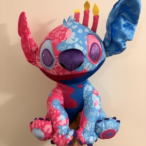 May include: A plush Stitch doll in pink and blue, with floral patterns and candle-like details on its head. The doll has large ears, purple eyes, and a purple nose. The doll is being held by a person.