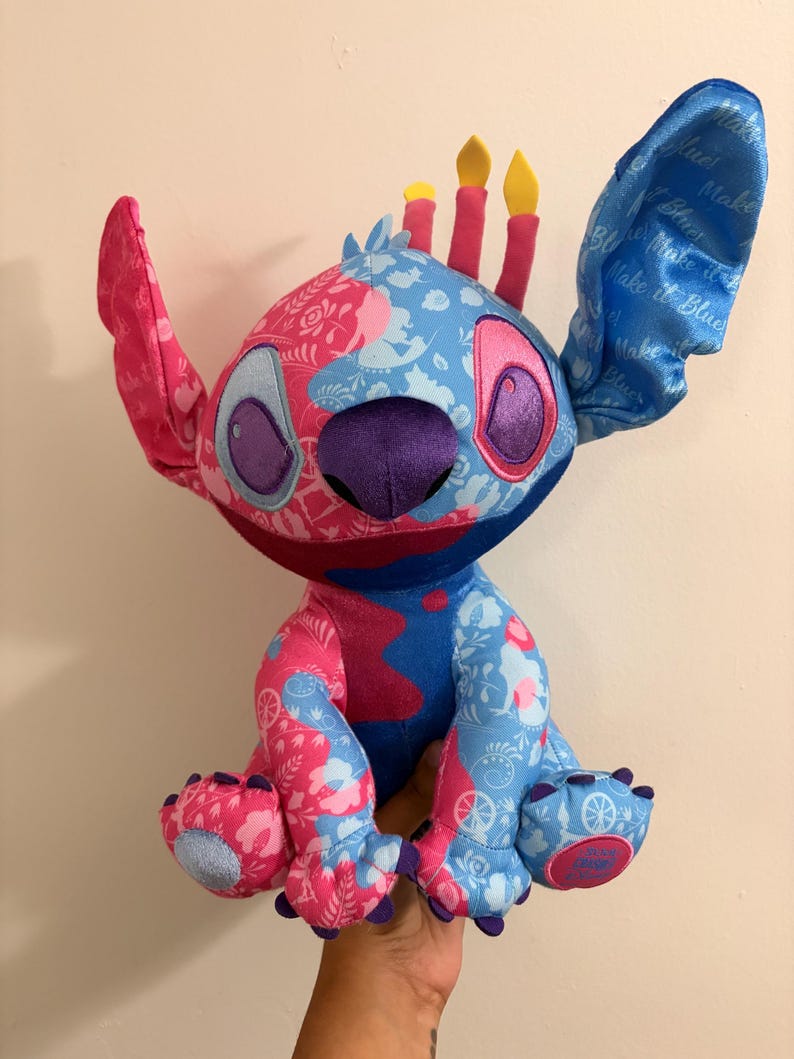 May include: A plush Stitch doll in pink and blue, with floral patterns and candle-like details on its head. The doll has large ears, purple eyes, and a purple nose. The doll is being held by a person.
