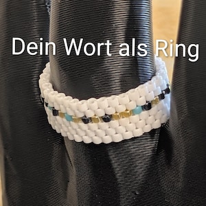 Könnte beinhalten: Ein weißer Perlenring mit goldenen, schwarzen und türkisfarbenen Akzenten. Der Ring wird auf einer schwarzen Oberfläche mit dem weißen Text "Dein Wort als Ring" präsentiert.