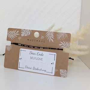 Könnte beinhalten: Ein schwarzes Schnurarmband mit kleinen Perlen auf einer braunen Papp-Präsentationskarte. Die Karte zeigt weiße Blattillustrationen, den Text MUSTHAVE und ein weißes Etikett mit dem Text Dein Code. SELFLOVE, Morsecode und Deine Bedeutung.