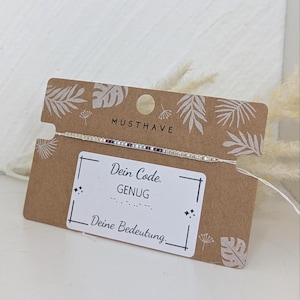 Könnte beinhalten: Ein dünnes Armband aus kleinen weißen und violetten Perlen auf einem hellen Faden, befestigt an einer braunen Papp-Präsentationskarte. Die Karte zeigt weiße Blattillustrationen, den Text MUSTHAVE und ein weißes Etikett mit dem Text Dein Code. GENUG.