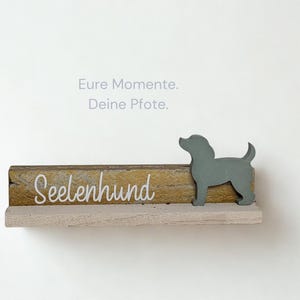 Könnte beinhalten: Ein Holzregal mit einer grauen Hundesilhouette und dem Wort "Seelenhund" in weißer Schrift. Das Regal hat eine hellgraue Basis und eine goldfarbene Holzoberfläche. Der Text "Eure Momente. Deine Pfote." steht darüber.