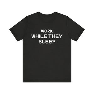 May include: Black t-shirt with the white text "WORK WHILE THEY SLEEP" in a bold, sans-serif font. The shirt is a crew neck style, and the text is centered on the chest.