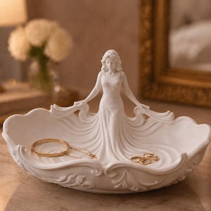 May include: A white ceramic jewellery dish featuring a sculpted woman in a flowing gown. The dish holds a gold bracelet, necklace, and rings. The dish has an ornate, scalloped edge.