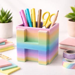 Könnte beinhalten: Ein pastellfarbener Schreibtisch-Organizer in Regenbogenfarben in Form des Buchstabens "H" enthält Stifte, Scheren und einen Bleistift. Der Organizer ist von Haftnotizen, Washi-Tape und kleinen Topfpflanzen umgeben und schafft so einen farbenfrohen und organisierten Arbeitsplatz.