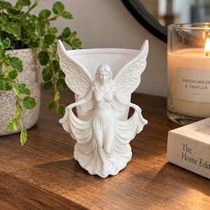 May include: A white ceramic planter shaped like a winged figure with flowing robes. The figure stands with arms outstretched, holding the edges of a draped garment that forms the vessel. The surface is matte white with detailed sculpted wings.