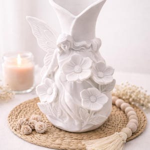 May include: White ceramic vase with a fairy design, featuring a fairy with wings and floral accents. The vase has a flared top and is decorated with sculpted flowers. A candle and decorative beads are in the background.