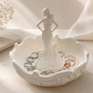 Peut inclure: Un vide-poche en céramique blanche avec une figurine centrale représentant une silhouette debout, les mains sur les hanches. Le bol circulaire présente un bord festonné et contient plusieurs bagues argentées et un collier de perles blanches.