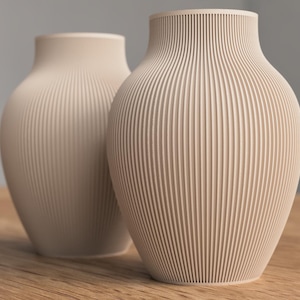 Op de afbeelding: Twee beige keramische vazen met verticale geribbelde details. De vazen hebben een afgeronde vorm met een licht uitlopende opening. Ze worden gepresenteerd op een houten oppervlak tegen een neutrale achtergrond. De vazen zijn qua grootte en ontwerp vergelijkbaar.
