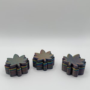 May include: Three stacks of colorful, leaf-shaped coasters. Each coaster is made of layered material with a rainbow effect, showcasing shades of blue, green, purple, and gold. The coasters are arranged on a white surface.