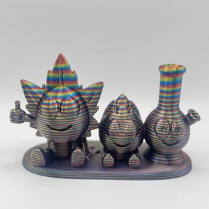 May include: A set of three whimsical, rainbow-colored figurines. The set includes a smiling character with a cannabis leaf head, a smiling egg-shaped character, and a smiling bong. All three are on a matching base.