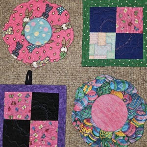 Puede incluir: Cuatro salvamanteles acolchados de colores. Dos tienen forma de flor, uno con conejitos y otro con huevos de Pascua. Los otros dos son cuadrados, uno con borde verde y otro con borde morado. Cada uno tiene un lazo negro para colgar.