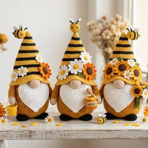 May include: Three handmade crochet gnomes with yellow and black striped hats, white beards, and sunflower and daisy accents. Each gnome has a different accessory, including a bee, a honey pot, and a bouquet of flowers. The gnomes are on a white wooden surface.