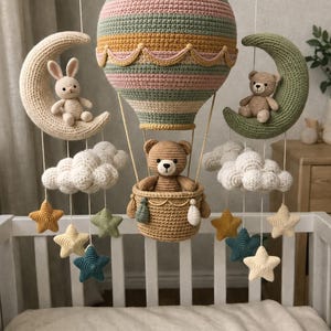 Puede incluir: Móvil de bebé hecho a mano con ganchillo, con un globo aerostático, osos de peluche, un conejito, nubes, lunas y estrellas. El globo tiene rayas de colores pastel. El móvil cuelga sobre una cuna blanca.