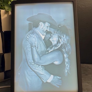 May include: A rectangular, black-framed light box displaying a monochromatic, backlit lithophane image of two people embracing. The person on the left wears a cowboy hat and a suit jacket, while the person on the right has long hair.