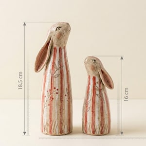May include: Two ceramic rabbit figurines. The larger one is 18.5 cm (7.3 inches) tall, and the smaller one is 16 cm (6.3 inches) tall. Both have a beige base with red and white vertical stripes and floral accents.