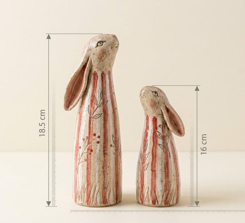 May include: Two ceramic rabbit figurines. The larger one is 18.5 cm (7.3 inches) tall, and the smaller one is 16 cm (6.3 inches) tall. Both have a beige base with red and white vertical stripes and floral accents.