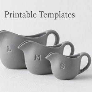 Op de afbeelding: Drie grijze keramische kannen van verschillende formaten, gemarkeerd met L, M en S, met handvatten en tuiten. De tekst "Printable Templates" staat boven de kannen.