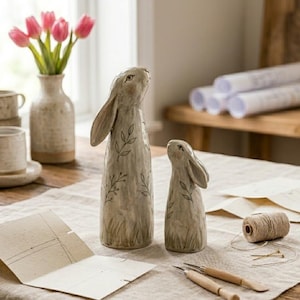 May include: Two ceramic rabbit figurines with leaf and stem designs. The rabbits are light beige with long ears and are looking upwards. A vase of pink tulips and a cup and saucer set are in the background.
