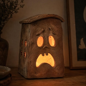 May include: A brown, textured ceramic lantern shaped like a house with a sad, expressive face. The structure features cut-out oval eyes and a wide, downturned mouth, with smaller square windows on the side. A warm, glowing light emanates from within.
