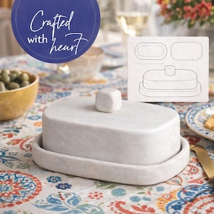 May include: A white marble butter dish with a square knob on the lid. The dish sits on a matching base, and the text "Crafted with heart" is in a blue circle in the background. The dish is on a colorful patterned tablecloth.