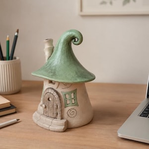 May include: A ceramic decorative house figurine featuring a cream-colored base with a textured arched wooden door, a small green window, and a prominent, curved, sage green roof with a chimney. The structure has a whimsical, rounded shape.